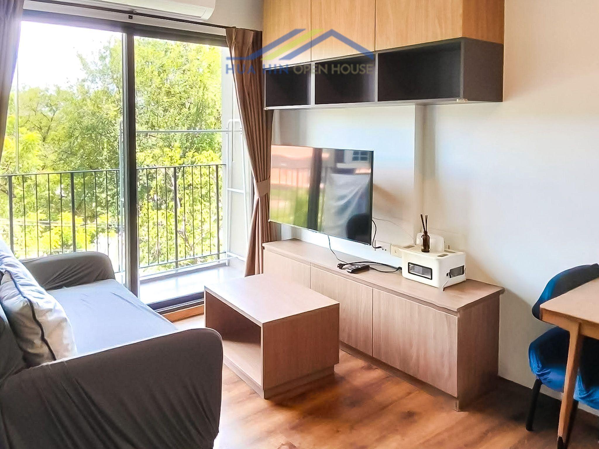 Modern living area with a sofa, TV, and balcony access, surrounded by greenery in a 1-bedroom condo.