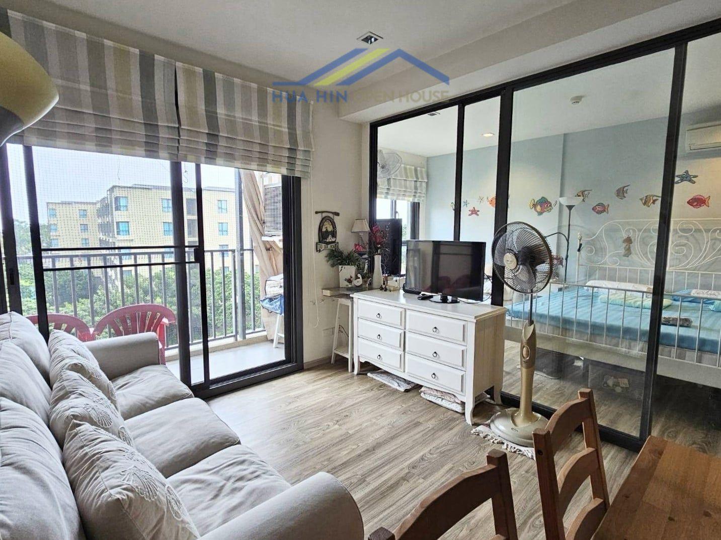 Rain Cha-Am living area with a sofa, dining table, and glass doors leading to a balcony; bedroom visible through glass partition.
