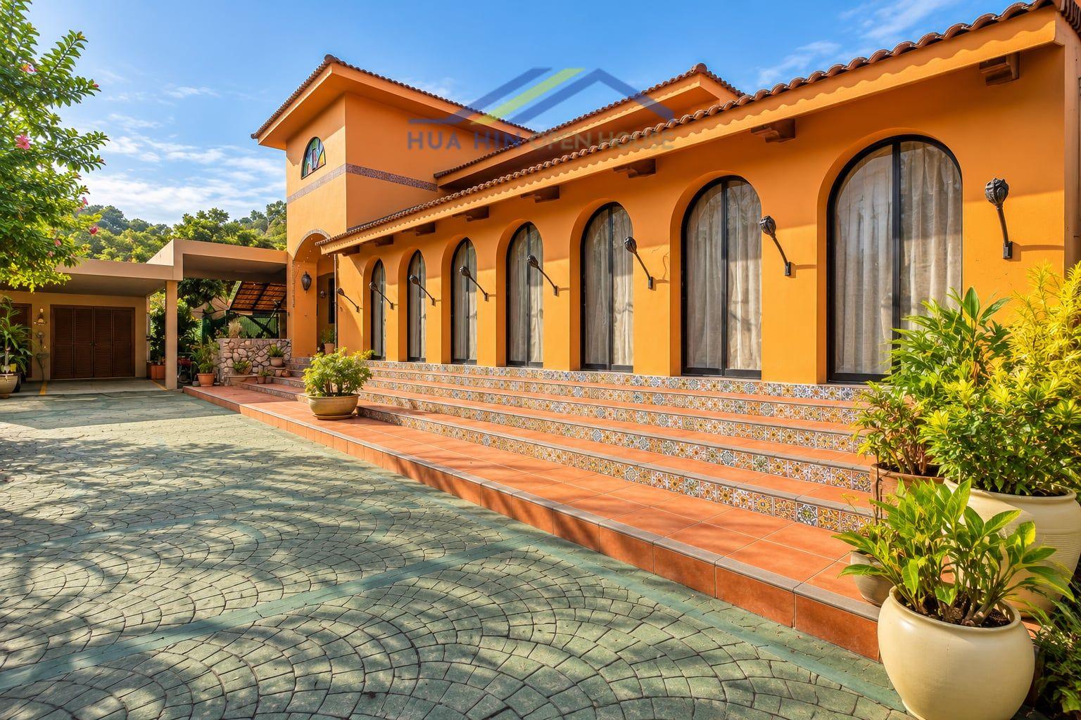 Hacienda-style villa exterior featuring arched windows, vibrant orange walls, and lush landscaping in Hua Hin.