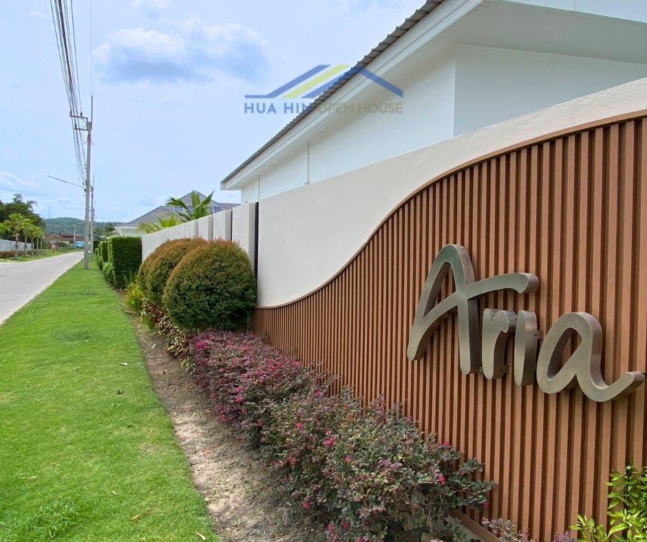 Modern entrance sign for Aria Hua Hin, surrounded by landscaped greenery and colorful plants along the pathway.
