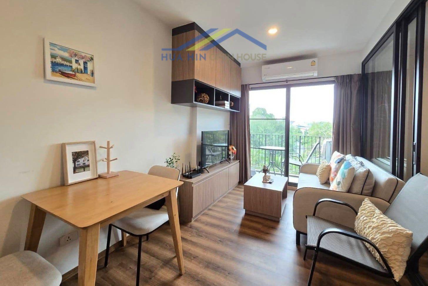 Modern living area in a 1-bedroom for Rent at La Habana Hua Hin, featuring a dining table, sofa, TV unit, and balcony access with greenery views.