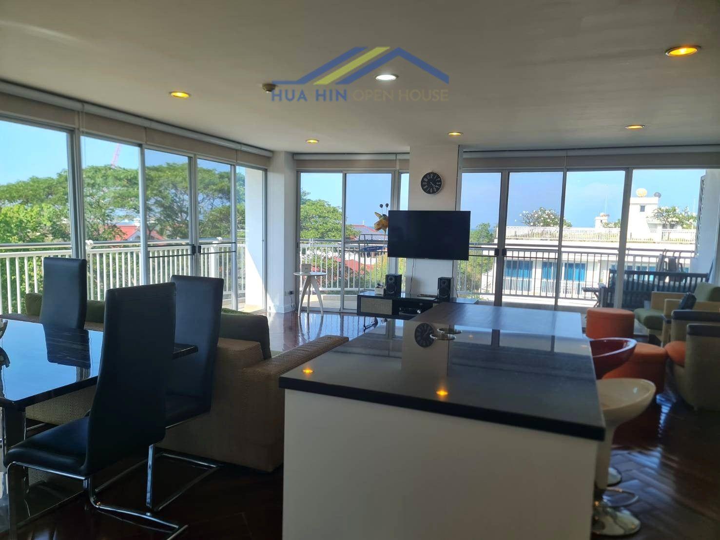 Spacious corner condo living area with large windows, modern furnishings, and a dining table overlooking greenery.