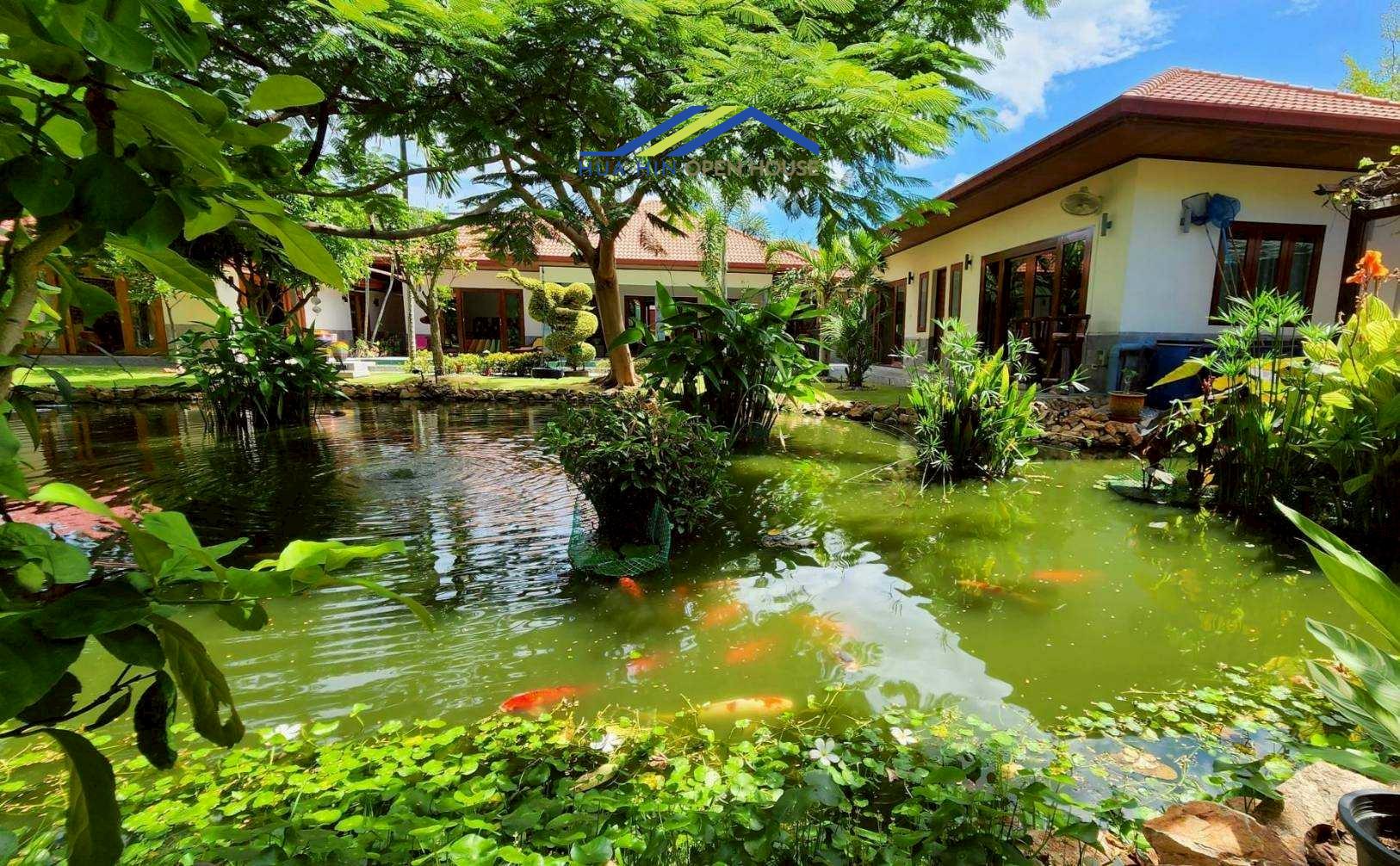Lush tropical garden with a serene pond, koi fish, and modern villa in the background, surrounded by greenery.